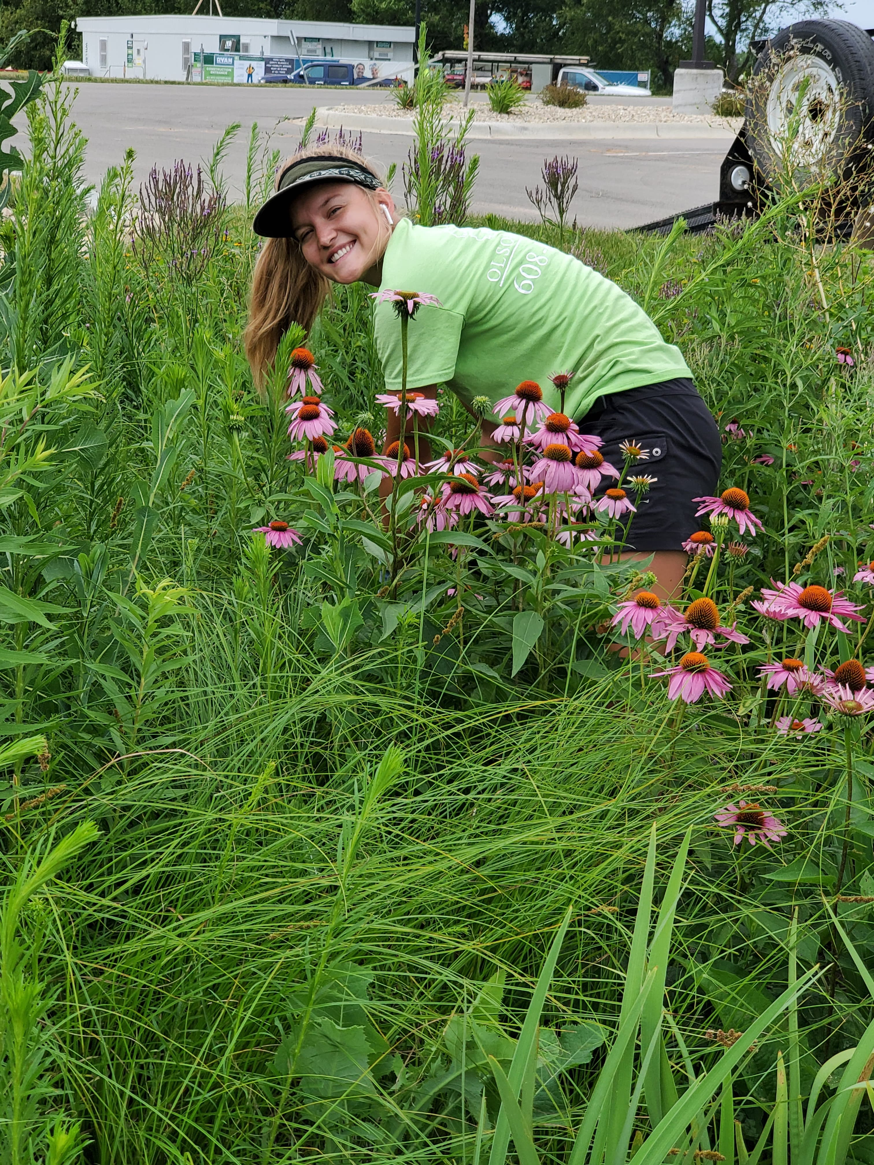 A smiling woman in a lime green shirt leans over purple coneflowers in tall grass.