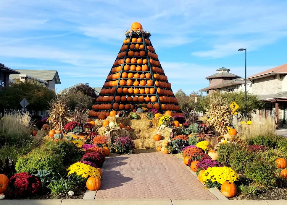 Tall pumpkin pyramid display surrounded by hay bales and colorful flowers on a sunny day.