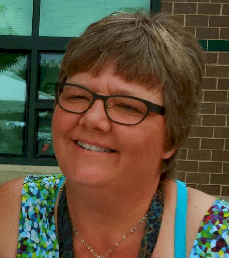 Smiling woman with short brown hair and glasses wearing a colorful, patterned sleeveless top.