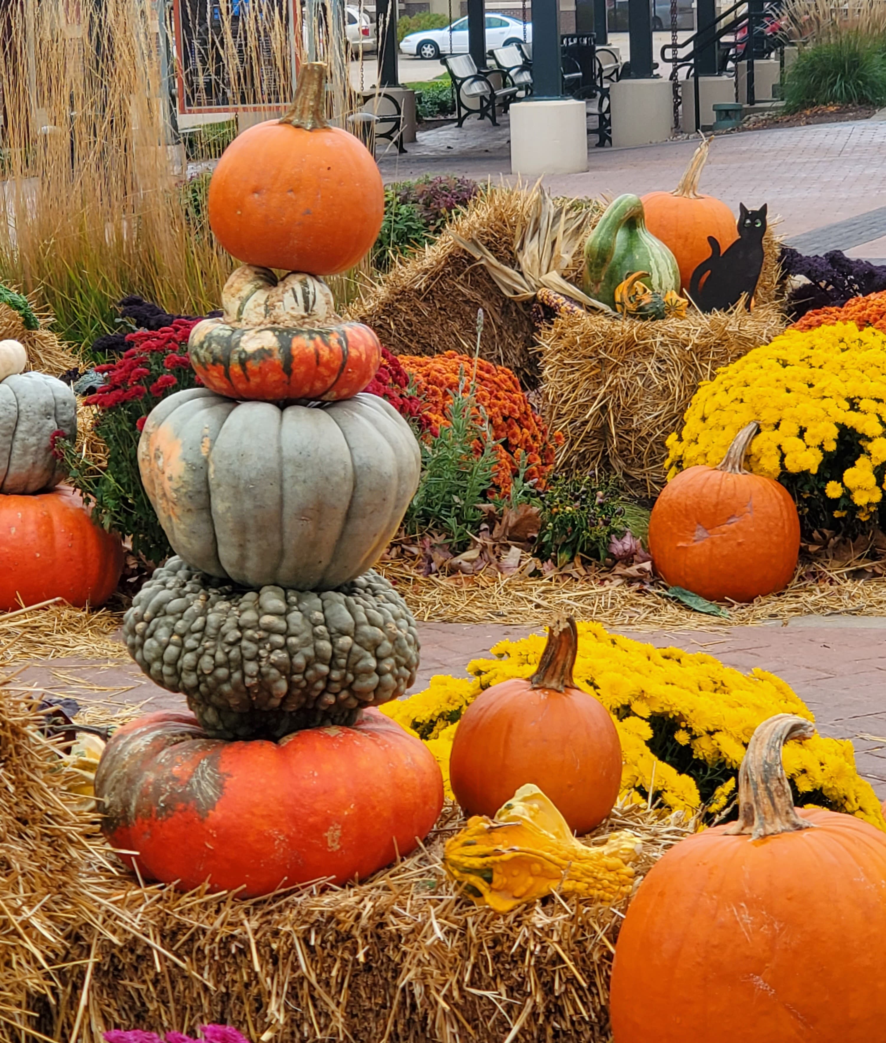 A tall stack of diverse pumpkins and gourds on hay bales with vibrant autumn flowers.